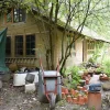 A photograph of a cabin in the woods. A wheelbarrow and plant pots sit out front.