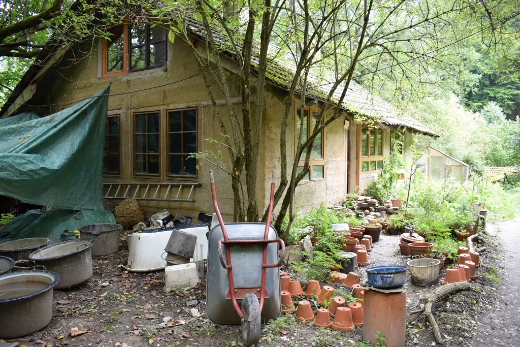 A photograph of a cabin in the woods. A wheelbarrow and plant pots sit out front.