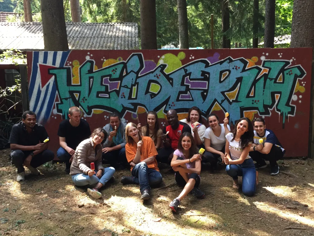 A photograph of volunteers smiling and eating popsicles in front of a sign which reads "Heideruh."